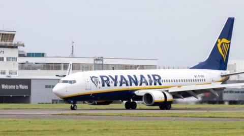 A Ryanair plane taxing at Cardiff Airport, with the terminal building in the background and a sign saying "Welcome to Wales" written at the left.