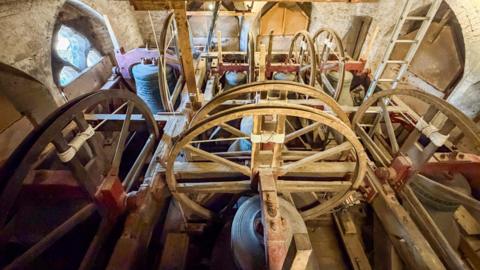 Church bells pictured from above in the tower. There are wooden wheels by each bell which are turned by rope from below in order to ring the bells.