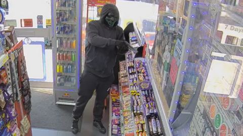 A man wearing a black hooded jacket, black trousers, black shoes, black gloves and a black horror-mask standi ng in a shop pointing what appears to be a black pistol. He is standing inbetween a rack filled with bags of sweets and a shelf filled with bars of chocolate.