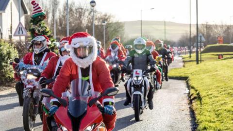 A convoy of bikers riding motorcycles down a narrow road while wearing Santa outfits, with a hill in the distant background and a building on the left side. 