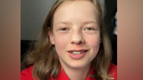 A teenage boy with brown eyes and shoulder length, dark blonde hair looks directly at the camera while smiling.  He is wearing a red tshirt.