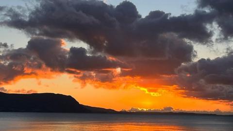 The sky is bright orange across the horizon as the sun sets behind a body of water, dark grey clouds are above