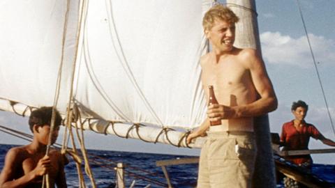 David Attenborough holds a bottle on a boat in a colourised image from Zoo Quest in 1956