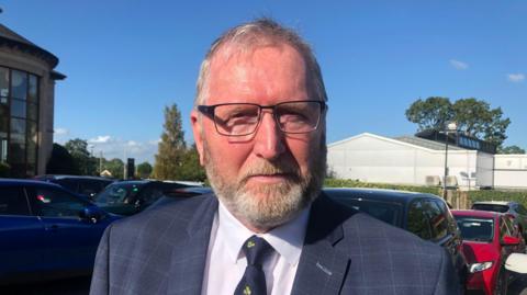 Doug Beattie has grey hair and a grey beard.
He is wearing wired framed, rectangular frames, a navy jacket, white shirt and navy tie. He's standing outside in a car park, a number of parked cars can be seen and there is a blue sky in the background.