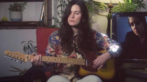 A woman with dark hair plays a yellow and brown guitar, while sat on a red chair in front of a white wall and some plants