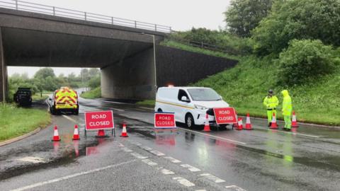Highways workers stand at the scene of a road closure following a helicopter crash near Okehampton, Devon. Three red ROAD CLOSED signs are on the road. There are several traffic cones lined up to block entrance and three vans parked along the road. The two men stood by the signs are wearing high-vis clothing. A bridge is running over the road.