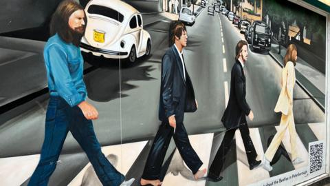 A tribute to the band Beatles - with four people painted walking on a crossroad on a road and cars can be seen parked in the background o either side of the road. One man is wearing a blue shirt and blue jeans, another is wearing a blackish suit, another is wearing a long coat and trousers and another man is wearing a cream suit.
