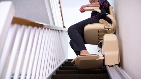 A woman sits in a stairlift which is going upwards. Only her legs and feet are visible. She is wearing blue trousers and sheer socks.