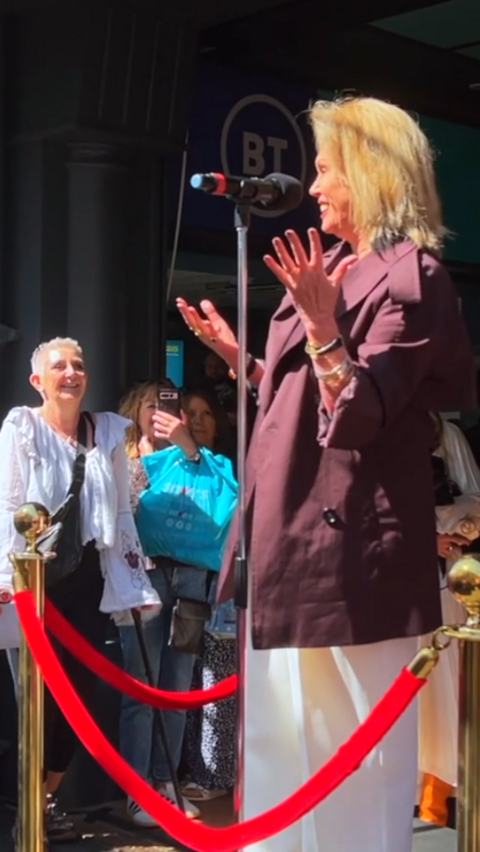 Joanna Lumley wearing white trousers and a burgundy jacket. She is standing on a platform behind red ropes, speaking into a microphone.