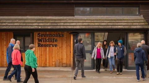 Visitors outside Sevenoaks Harrison Visitor Centre - it is a wood panelled building, with a tiled roof and a sign which has the name of the centre in black letters
