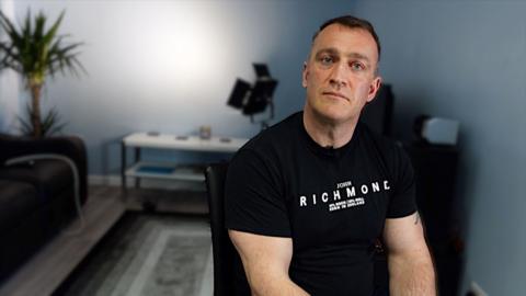 A well-built man in his late 30s sitting in a chair wearing a black t-shirt and looking at the camera. He is sitting in a grey-painted room with a back-lit wall and a plant in the corner. He is not smiling.