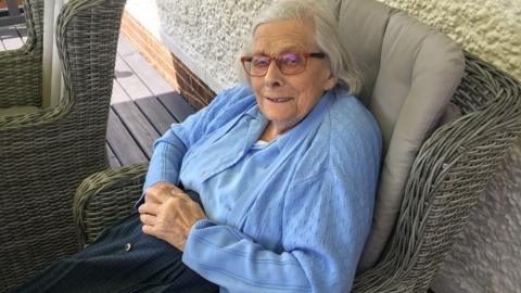 Elderly Valerie Knights sits in a garden chair and looks at the camera. She is smiling.