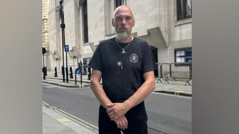 Piotr Kucharski standing outside the Central Criminal Court. He is wearing a black T-shirt and trousers and has a couple of necklaces. He is tanned with a shaved head and grey beard and moustache. 