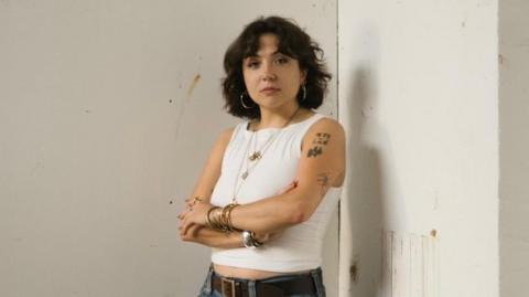 A woman with brown short hair wearing a white vest top, blue jeans with a brown belt. She is wearing bangles on both wrists and has tattoos on her left arm. 