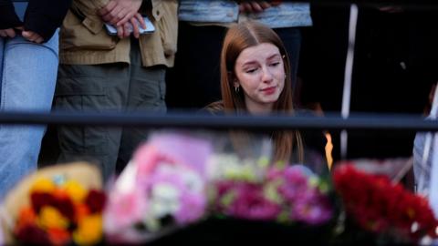 A young woman, with long, straight reddish brown hair, looks at floral tributes to Liam Payne with a sorrowful expression.