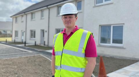 Steven Kemp is standing outside a housing development. He is wearing white hard hat and a high visibility vest over a pink polo shirt.