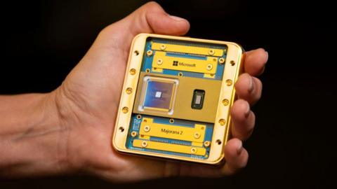 A hand holding the Majorana 2 chip in its palm. Including its housing the chip appears to be about the size of a beer mat. It consists of a gold plated metal frame and sits on a dark blue-green board bearing its name. The chip itself is a small silvery square on a dark gunmetal blue square with very fine gold circuitry visible. 