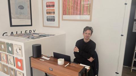 A person sits at a small wooden desk in a minimalist, gallery-like room with white walls. The person wears a dark long-sleeve top and sits with arms crossed, facing the camera. On the desk are a laptop, a white mug with a small pattern, and a pen. To the left, a display cabinet holds rows of vinyl records with colourful labels, and a stack of magazines or records is arranged on top. Several framed artworks hang on the wall behind, including designs featuring record labels and abstract patterns.