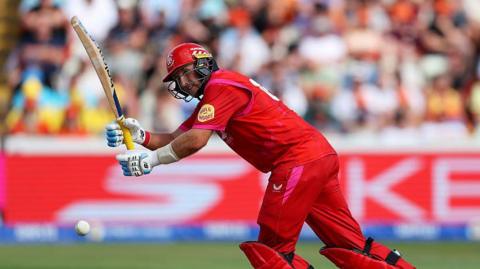 Joe Root, wearing red trousers, shirt and helmet, leans forward to play a shot with his bat held up in front of him