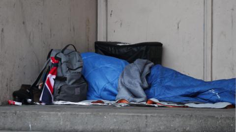 Person in a blue sleeping bag sleeping rough in a doorway with a union jack flag and a grey rucksack and empty cigarette packet.