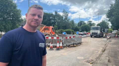 A man in a blue t-shirt with closely-cropped blond hair, stands in front of a set of roadworks. 