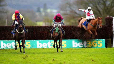 Three horses with their riders racing over a hurdle at Taunton race course. The jockeys are wearing bright yellow and pink tops.