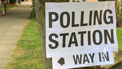 A sign saying "polling station, way in" is taped to a tree. In the background is a patch of green grass and a path to the left. Further up the path is another sign saying "polling station".