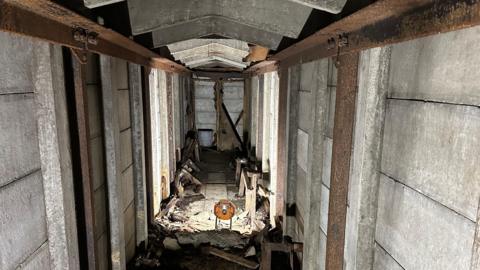 A concrete tunnel with metal support beams running along the walls. Debris can be seen on the floor.