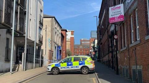 A police car parked across the street between two rows of buildings. A cordon can be seen behind the car, stretching across the street.