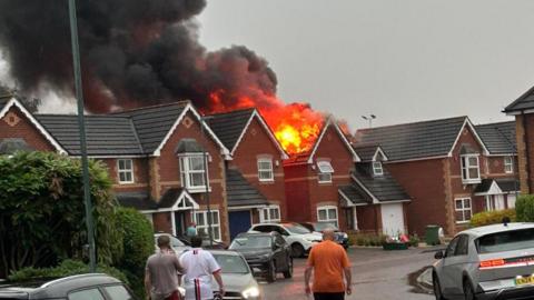 Fire erupting out of roof of detached house with cars and people in front.