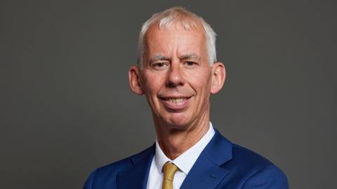 This is a close up photo of John Milne showing his head and shoulders. He is smiling. He has short white hair. He wears a navy blue suit, a white shirt and a gold tie. 