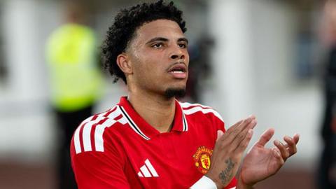 Ethan Williams in a red Manchester United shirt. He is clapping his hands to address fans in the crowd