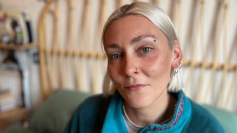 Elle Glenny, who has bobbed blond hair, looking directly at the camera and wearing a turquoise fleece. There is a bed frame in the background.