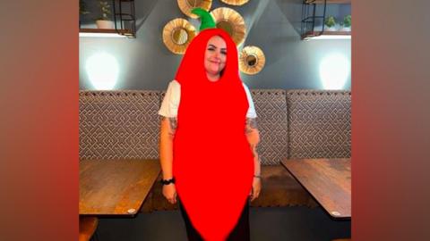 A woman smiling with a white t-shirt on and tattoo sleeve. She is dressed in a large red chilli costume which covers her head and hair and torso