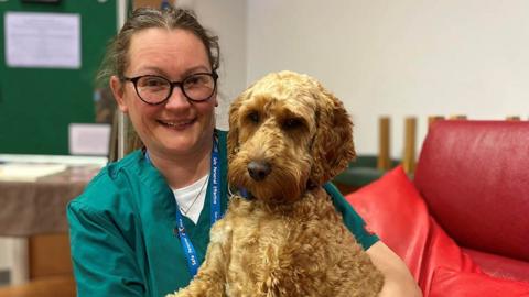 Rachel Fielding, who has tied-back dark hair and wears round dark-framed glasses, is photographed sitting on a red leather sofa. She is wearing green scrubs with her blue NHS lanyard around her neck. A golden brown cockerpoo dog called Alfie is sitting on her knee.