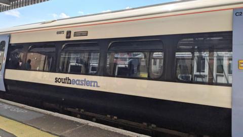 A white Southeastern train carriage, with blue doors, stopped at the platform with passengers inside, seen under the Margate Station canopy against a cloudy blue sky on a summer day in Kent
