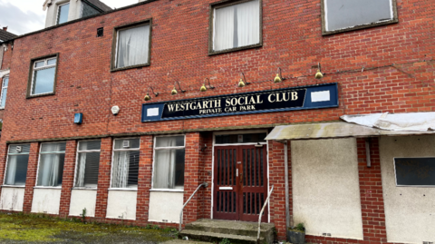 Exterior of Westgarth Social Club. It is a two-storey red bricked building with additional doormer windows in the roof. The windows on the ground floor are rectangular while the first floor ones are square. A long rectangular black sign above the central double doors gives the name of the venue and advertises a "private car park".