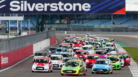 Dozens of Citroen C1 cars bunched together on the track at Silverstone circuit. The vehicles are of different colours including red, yellow, white and blue. The cars are passing a red and white barrier, behind which BRSCC banners are visible. There are empty spectator stands with blue sets behind the track.