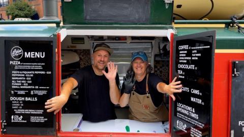 A man and woman are wearing black t-shirts and waving. They are holding open the shutters to a canal boat
