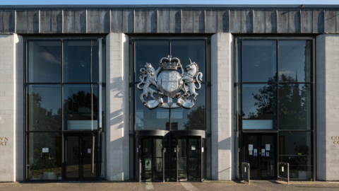 The outside of Coventry Crown Court, a glass-fronted building with grey stone pillars separated by large panels of glass. In the middle are two revolving doors and above them, the British coat of arms with a lion on one side of a shield, a unicorn on the right.