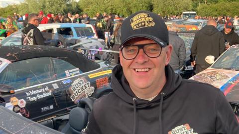 A man in a black hoody and black baseball cap surrounded by cars taking part in the challenge.