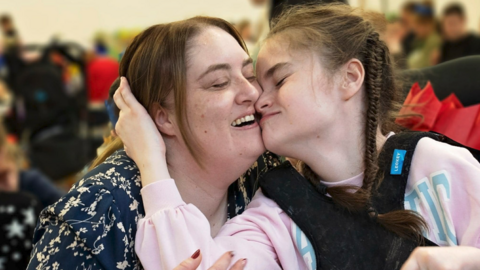 Claire and Kayla are nuzzling one another and Kayla has her hand around her mum's head and is smiling, as is her mother. Kayla has brown hair which is plaited . She has a pink jumper on and what could be a brace or strapping over her chest. Claire has a patterned top and brown hair.