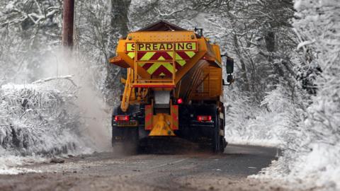 Orange gritting machine driving along snowy road, with snowy trees in the background.