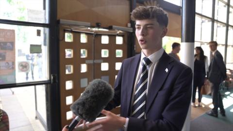 A young man with dark hair, wearing a blue suit and blue and white tie, speaks to a person holding a microphone