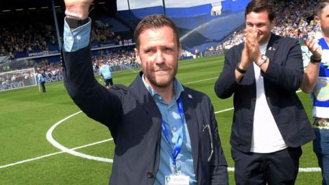 David Bruce is waving to the Sheffield Wednesday fans. He has short brown hair and is wearing a light blue shirt and dark jacket.