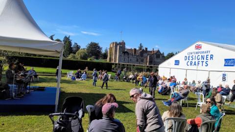People sat in plastic chairs on a lawn in the sun. A band is on a small stage under a gazebo and there is a large white tent with a sign that reads 'contemporary designer crafts'. A historic manor house can be seen behind a hedge.