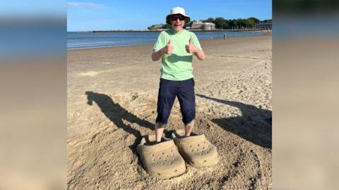 A man wearing a white hat, a light green t-shirt and blue cargo shorts, standing on a beach, smiling and giving a double thumbs up. He's standing in a pair of giant crocs that have been carved out of the sand.