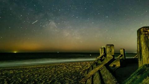 night sky image from a beach overlooking the sea. Lots of bright white spots in the sky and one streak of light showing a meteor streak