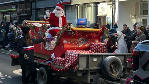 A trailer being tower through a street with people watching. Father Christmas is on top, waving. He is wearing a red outfit with white fur and a red and white hat. He has a long white curly beard. The trailer has been turned into a red and gold sleigh, with wrapping paper around it.