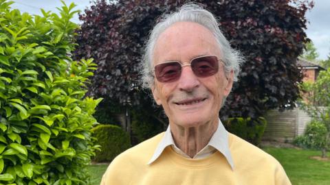 An older man with grey hair and dark glasses facing the camera as he stands in a garden with a lawn, bushes, trees and flower beds in the background. He is wearing a white shirt and a yellow top which has a footprint on it with the words "a nineties Odyssey" written in the middle.